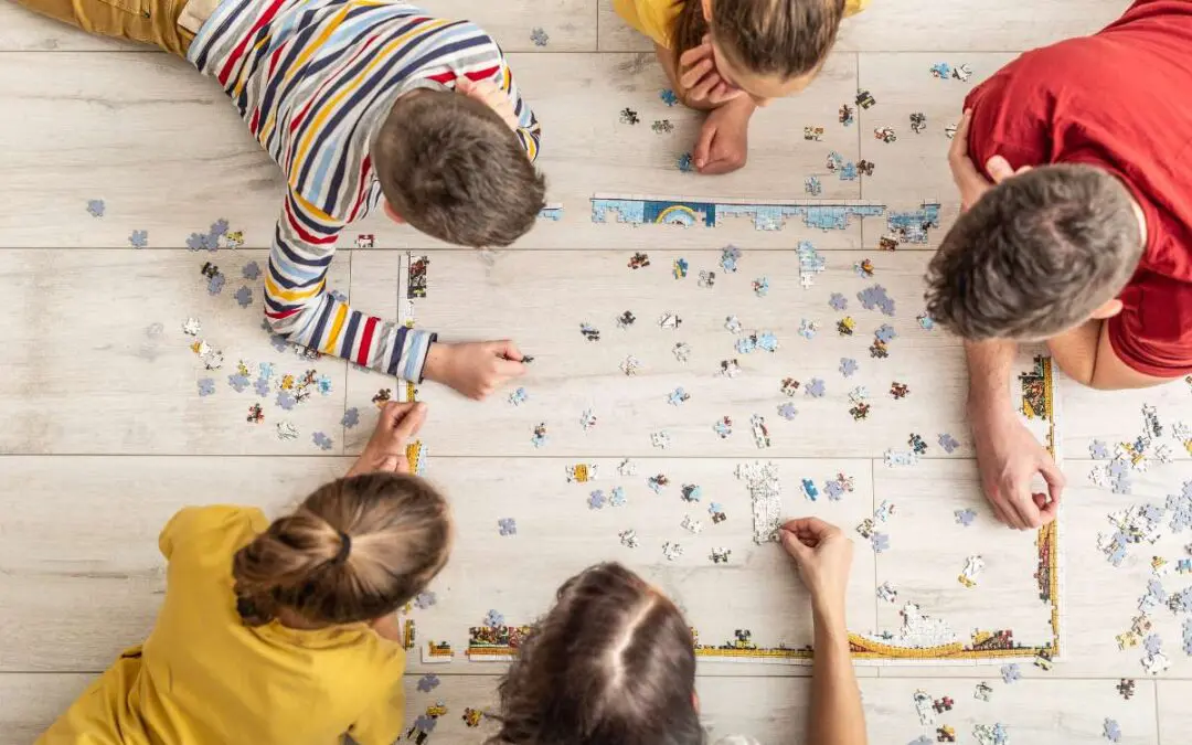 A family lies on the floor working together to assemble a large jigsaw puzzle.