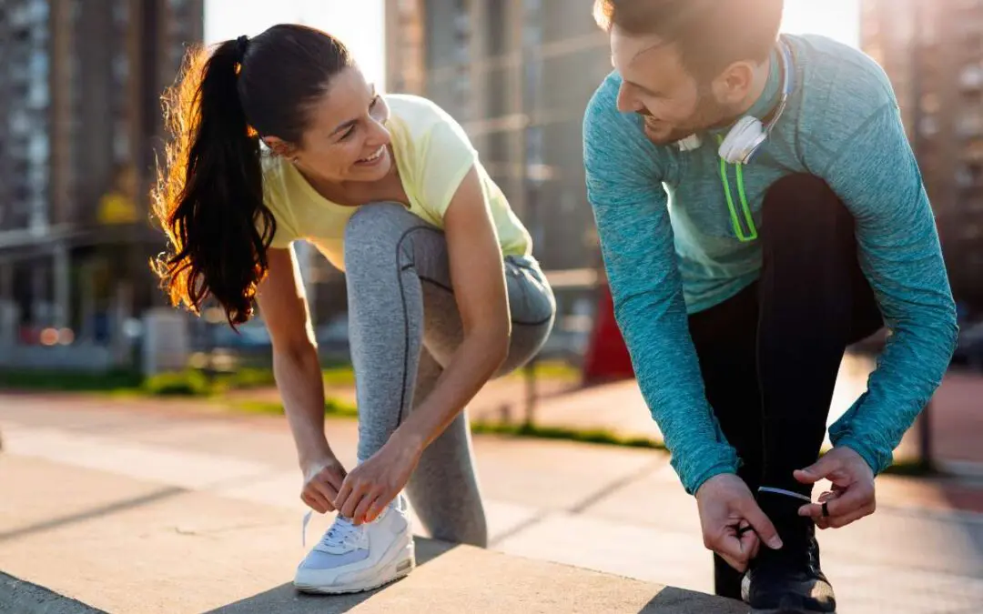 A man and woman smile at each other while tying their sneakers before exercising outdoors.