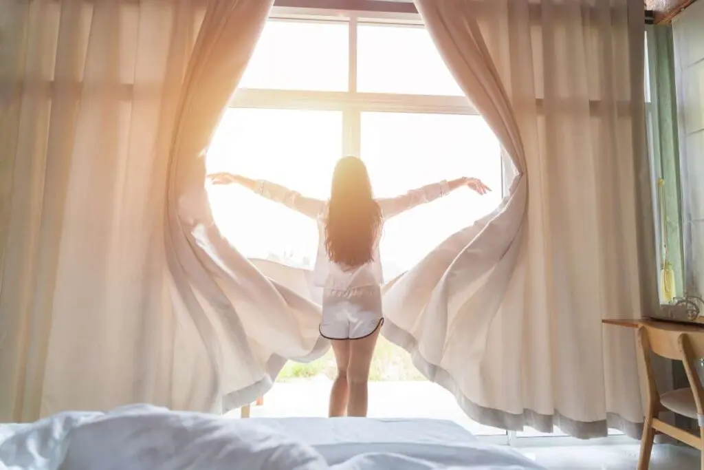 A girl opens wide curtains in the morning, letting sunlight flood into the bedroom.