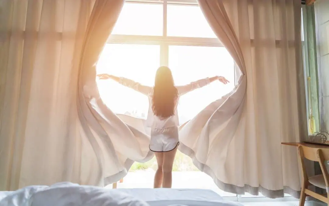 A girl opens wide curtains in the morning, letting sunlight flood into the bedroom.