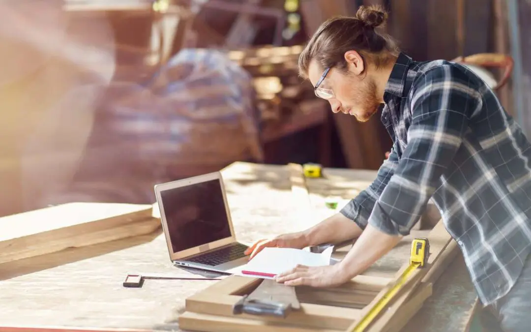 A man in a workshop leans over plans and his laptop while working with wood.