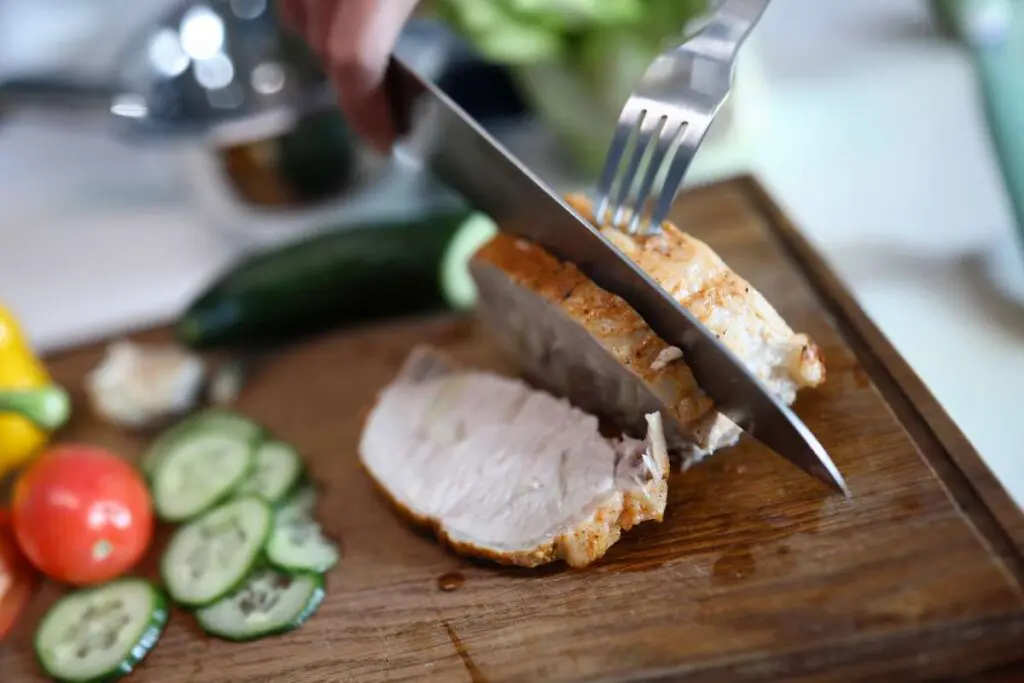 A person cutting meat using knife and fork on a wooden board.