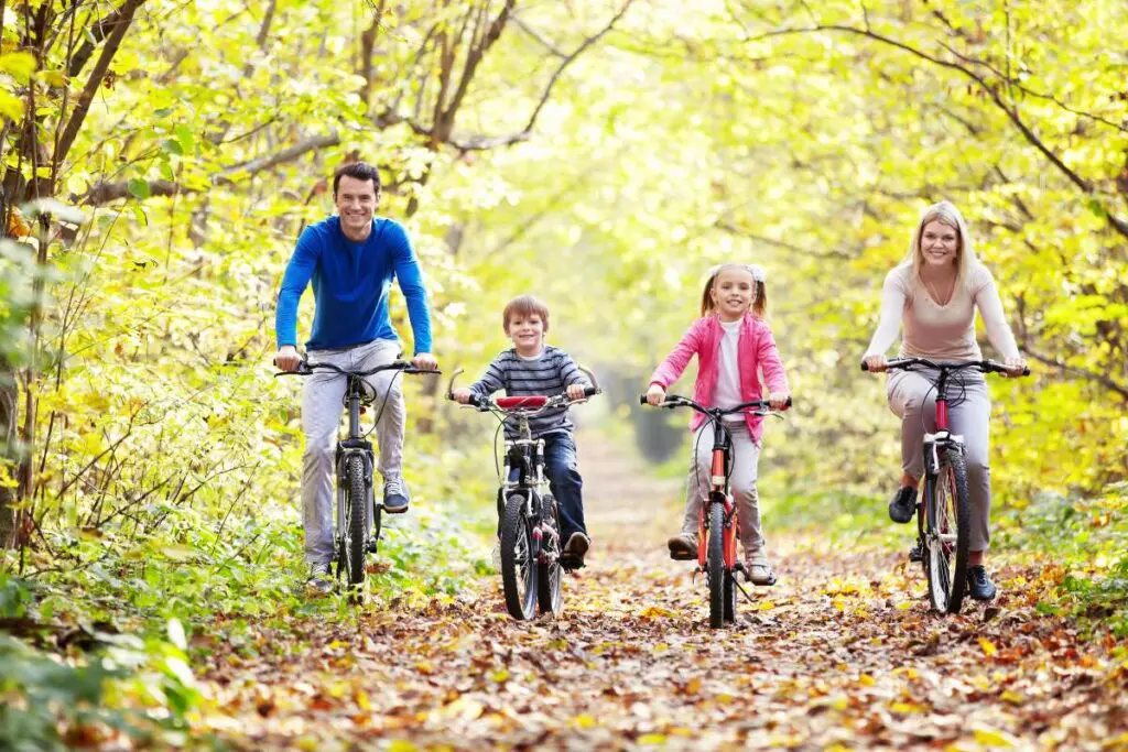A family of four is riding bicycles together on a leaf-covered path.