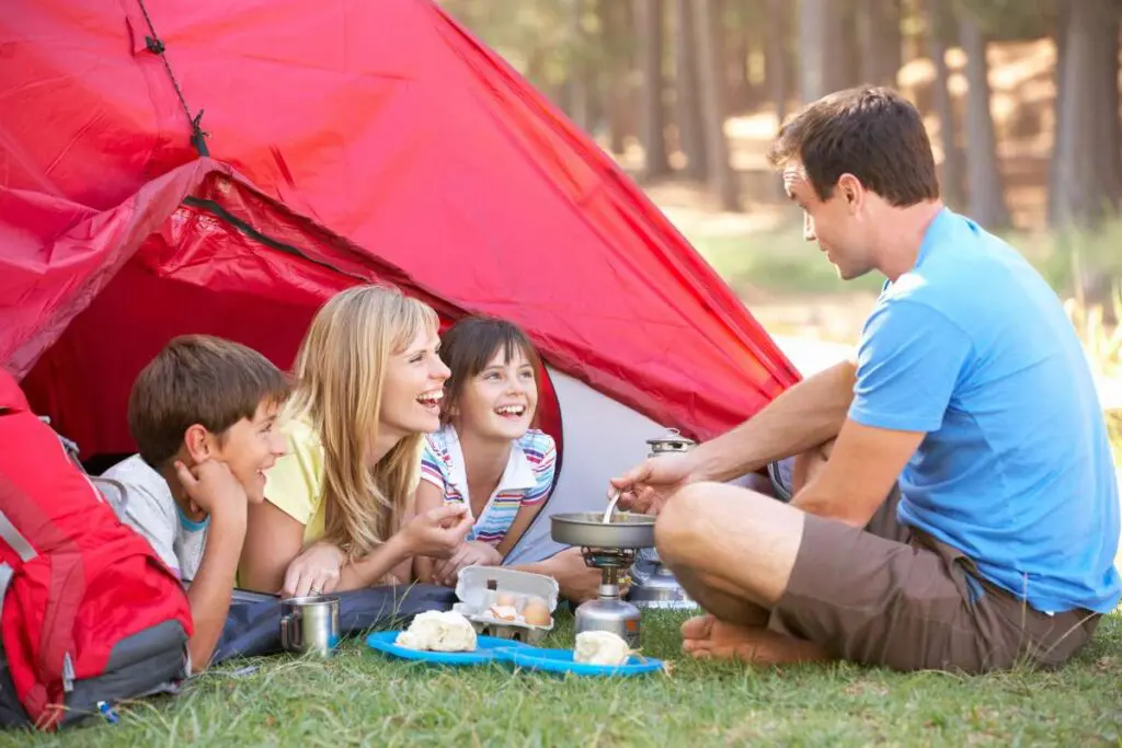 A family is happily camping in a red tent while their father is cooking.
