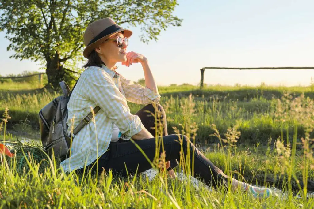 Woman in hat with backpack sitting on the grass enjoying summer nature, rural landscape, golden hour sunset.