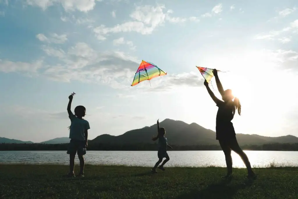 Children are flying kites while running on a meadow by the lake at sunset with their mother.