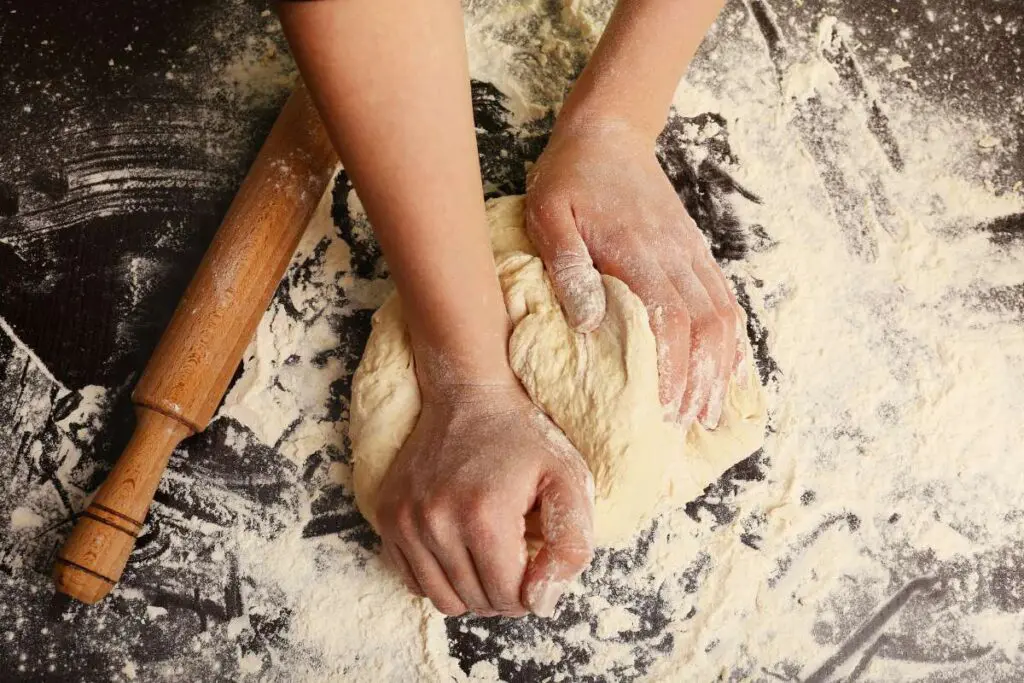 A hands making dough on wooden table; a wooden roller is next to it.