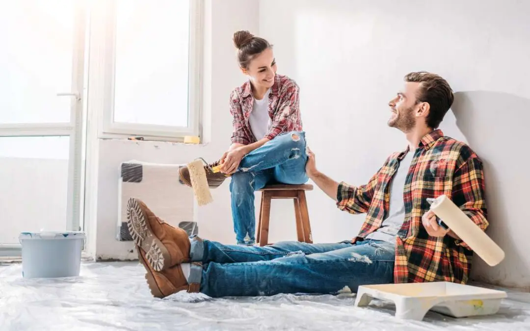 A couple sitting while holding paint rollers and smiling each other.