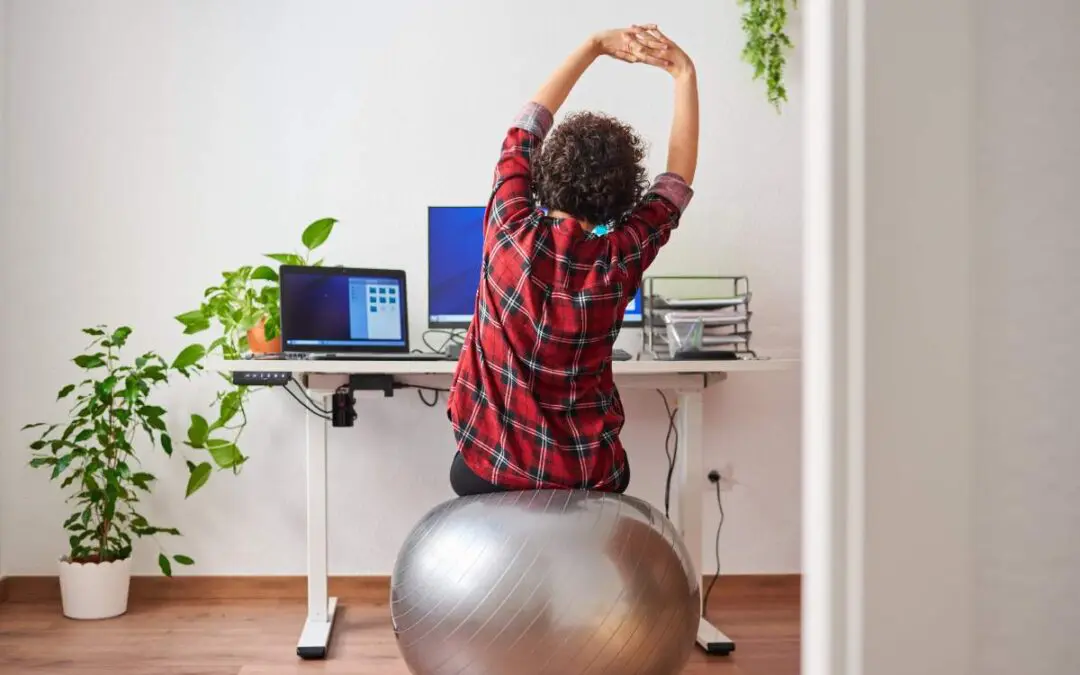 A woman is stretching in front of her work desk while sitting on a fit ball.
