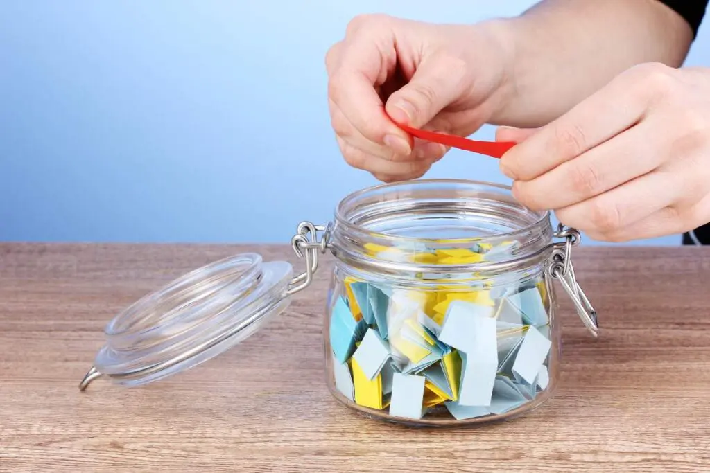 A person holding a red piece of paper above a glass jar filled with folded papers on a wooden table.