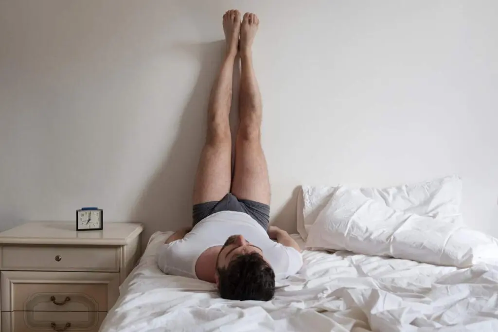 A man is lying in bed with his legs up on the wall.