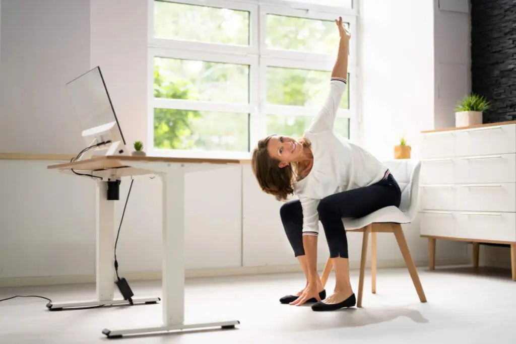 A woman is sitting on a chair beside a desk, stretching her body sideways with one arm up and the other on the floor.