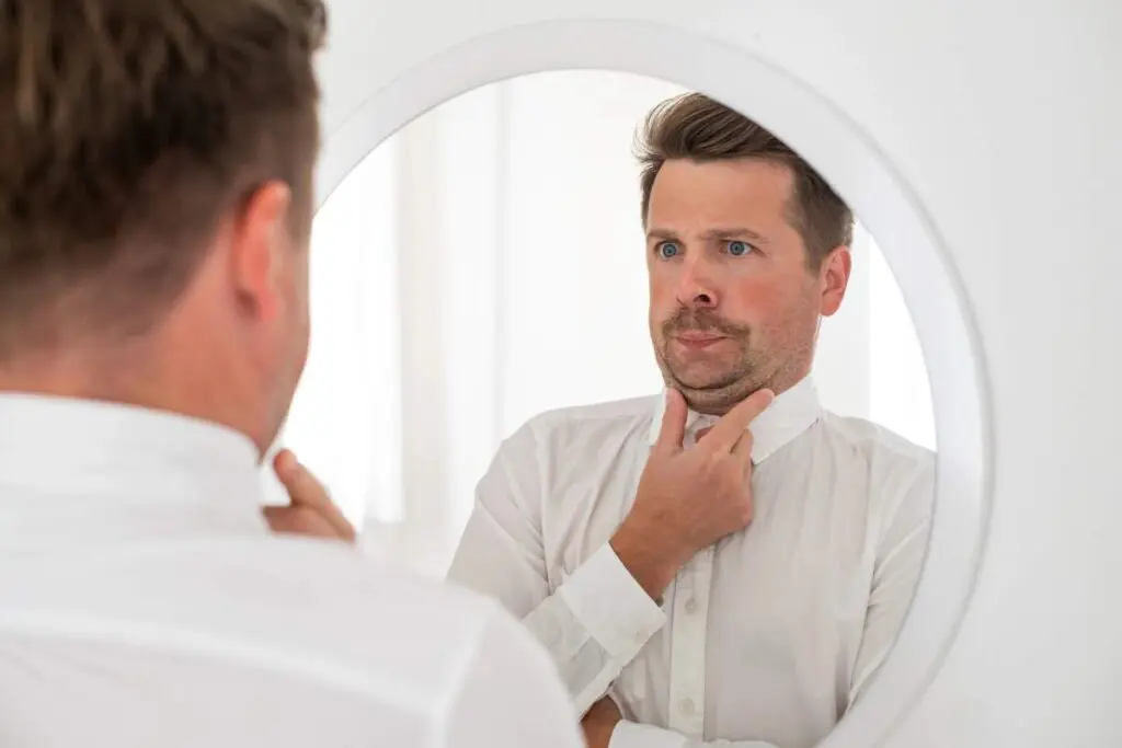 A man is standing in front of a mirror while pressing his chin down toward his neck.