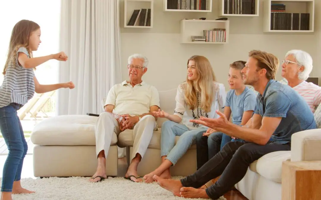 A young girl performs a charades gesture while her family sits on the couch watching.