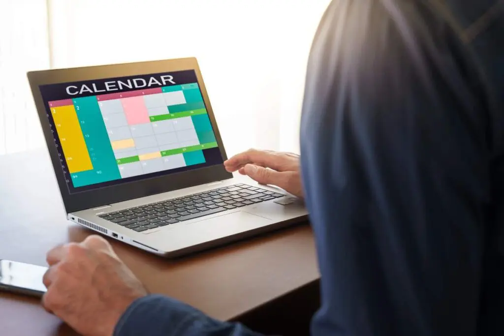 A person sits at a desk using a laptop with a colorful digital calendar displayed on the screen to organize their schedule.