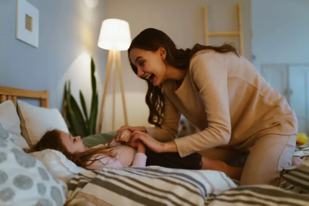 A mother is kneeling on the bed smiling as she tucks her daughter in bed.