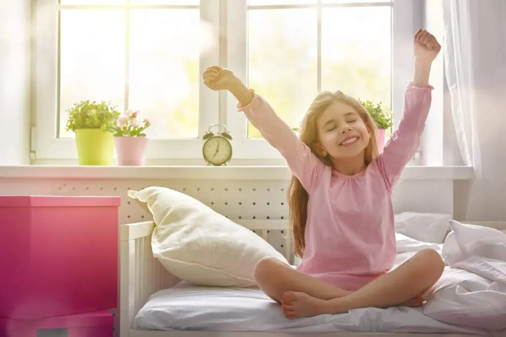A little girl is stretching while sitting on bed on a bright day.