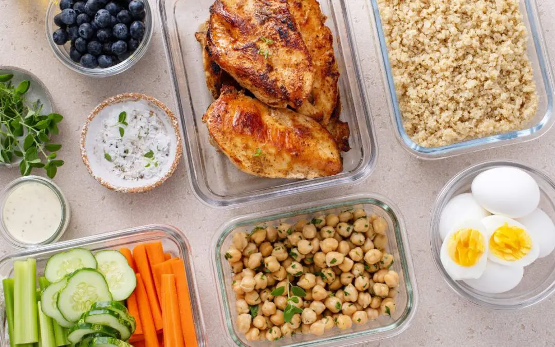 Glass containers are filled with grilled chicken, chickpeas, quinoa, eggs, and fresh vegetables.