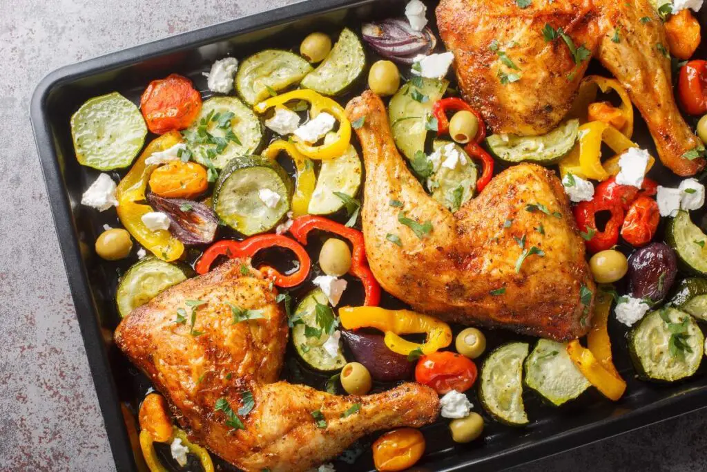 A tray holds roasted chicken legs surrounded by colorful vegetables, olives, crumbled cheese, and herbs.