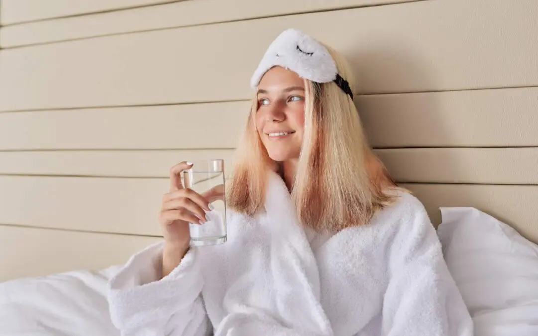 A woman wearing a bathrobe and sleeping mask is in bed holding a glass of water