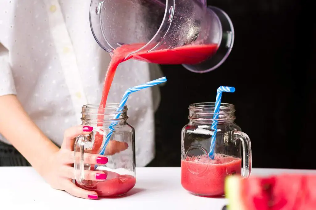 A person pours watermelon smoothie from a blender into a glass jar with a blue straw.