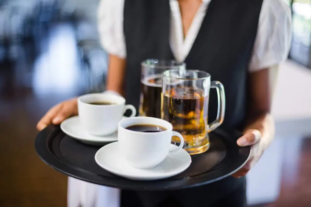 A server carries a tray holding two cups of black coffee and two glass mugs of beer.