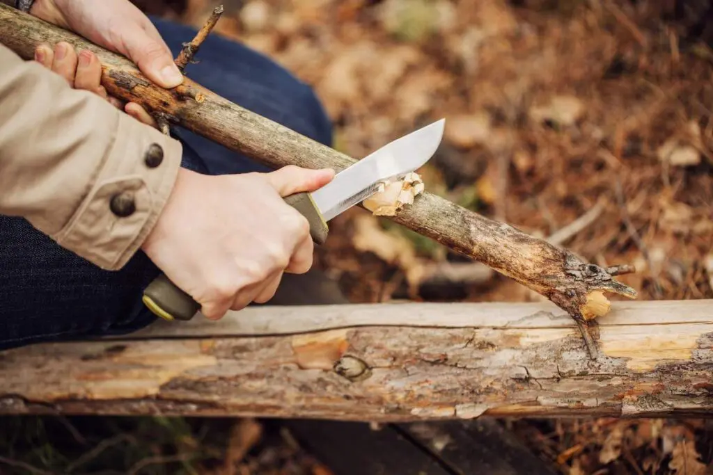 A person uses a knife to carve or shave a branch outdoors.