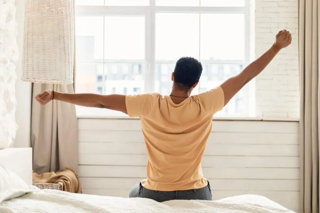 A man sitting on a bed, arms raised, in an indoor room with a window and curtains.