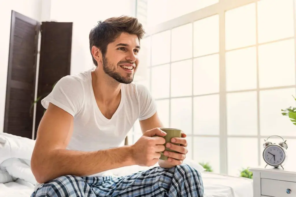 A man sitting on a bed, holding a cup, with a window and vase in the background.