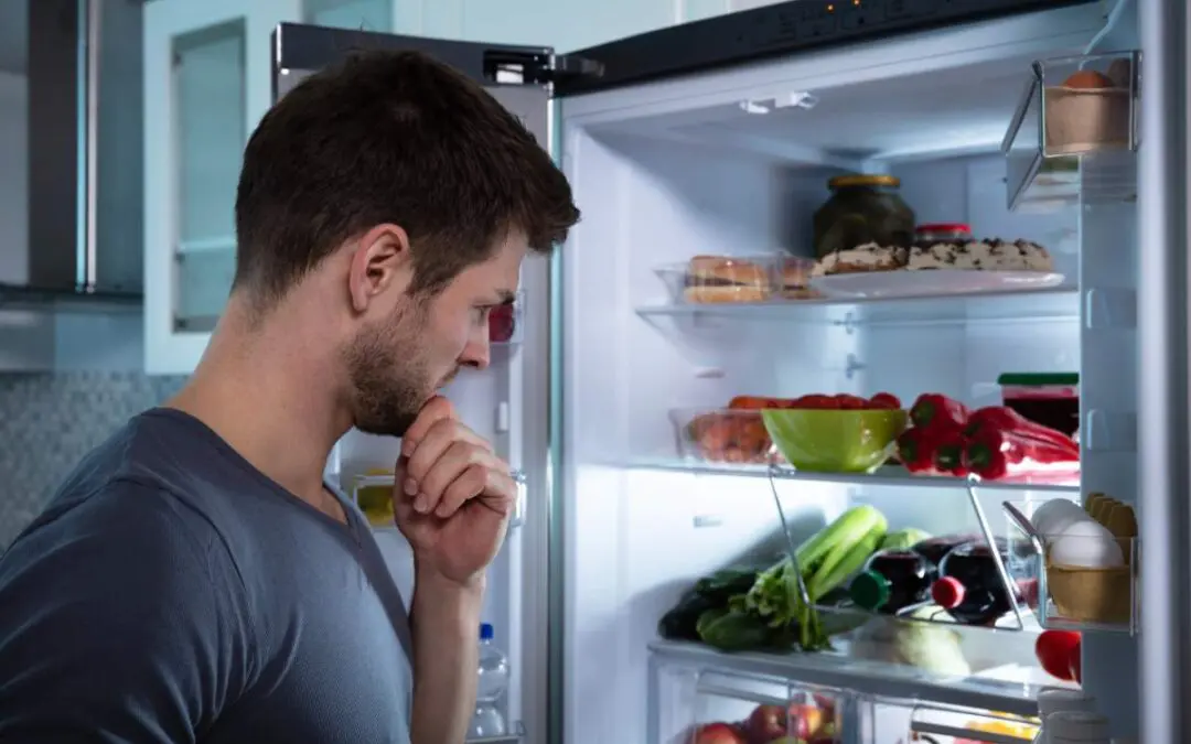 A man is standing in a kitchen, looking at an open refrigerator filled with food.