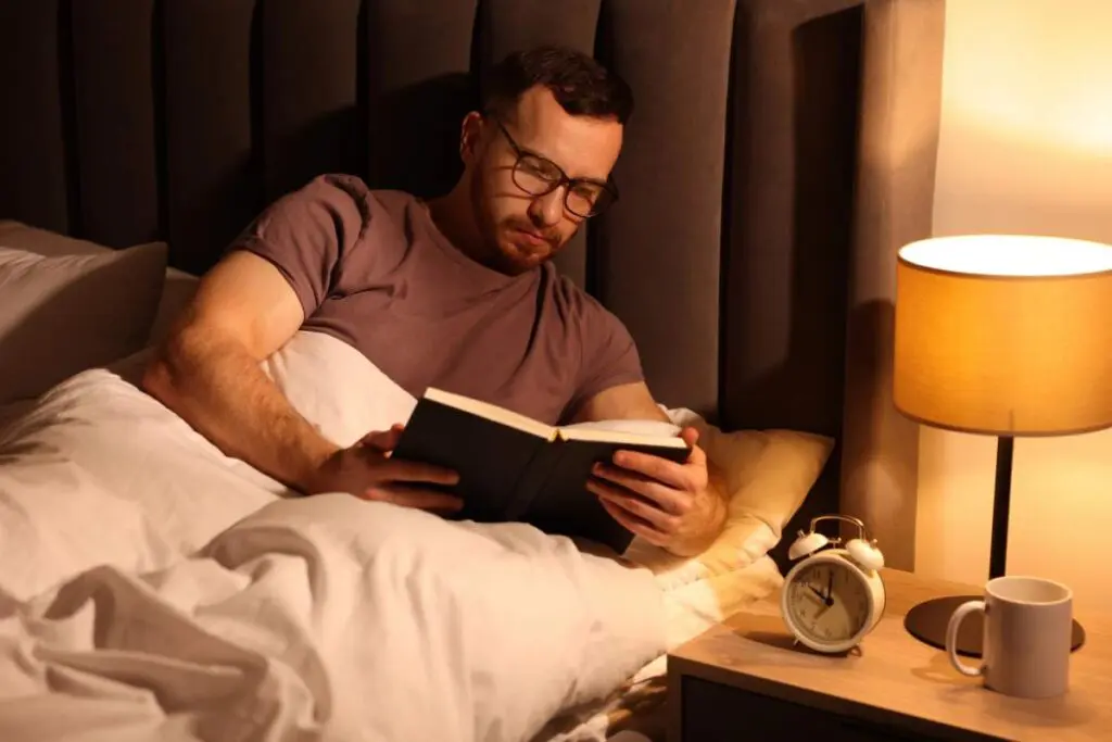 A man lying in bed reading a book, with a wall and lamp visible in the background.