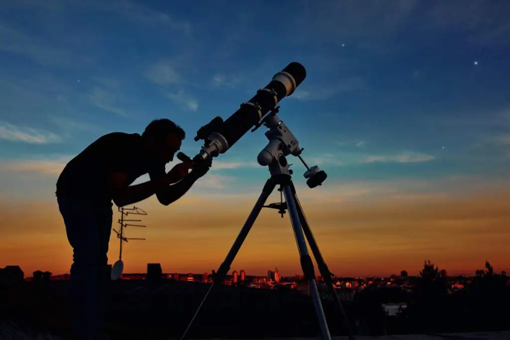 A man observing the sky through a telescope mounted on a tripod during sunset.
