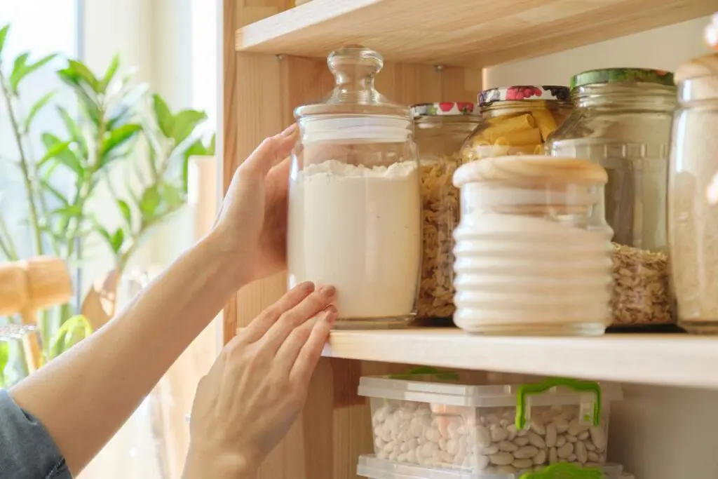 A person reaching for a jar of flour on a shelf in a pantry filled with various containers and jars.