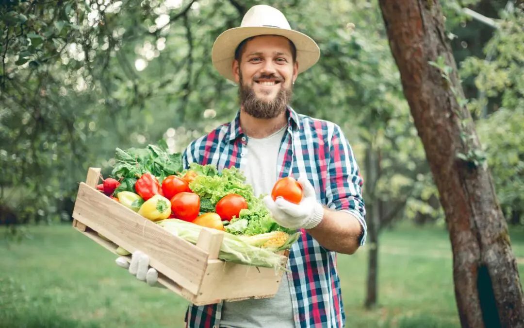 A man wearing a straw hat and gloves is smiling while holding a basket filled with fresh vegetables.