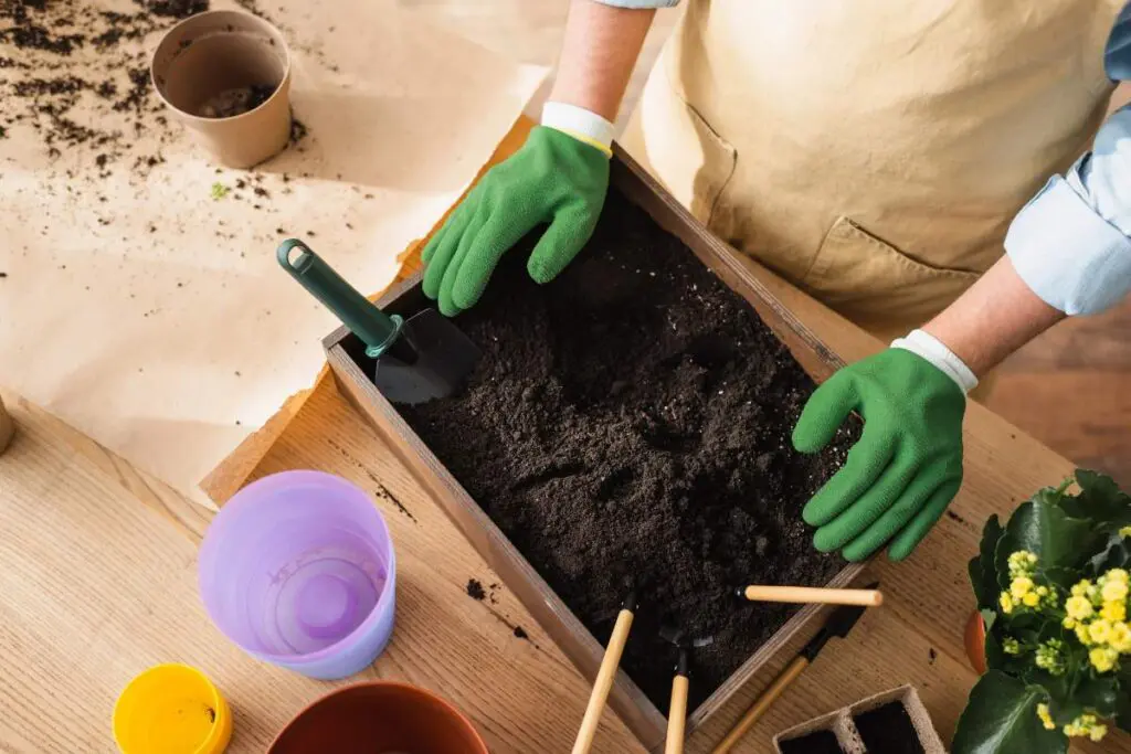 A person wearing green gardening gloves is using a trowel in a wooden planter filled with soil.