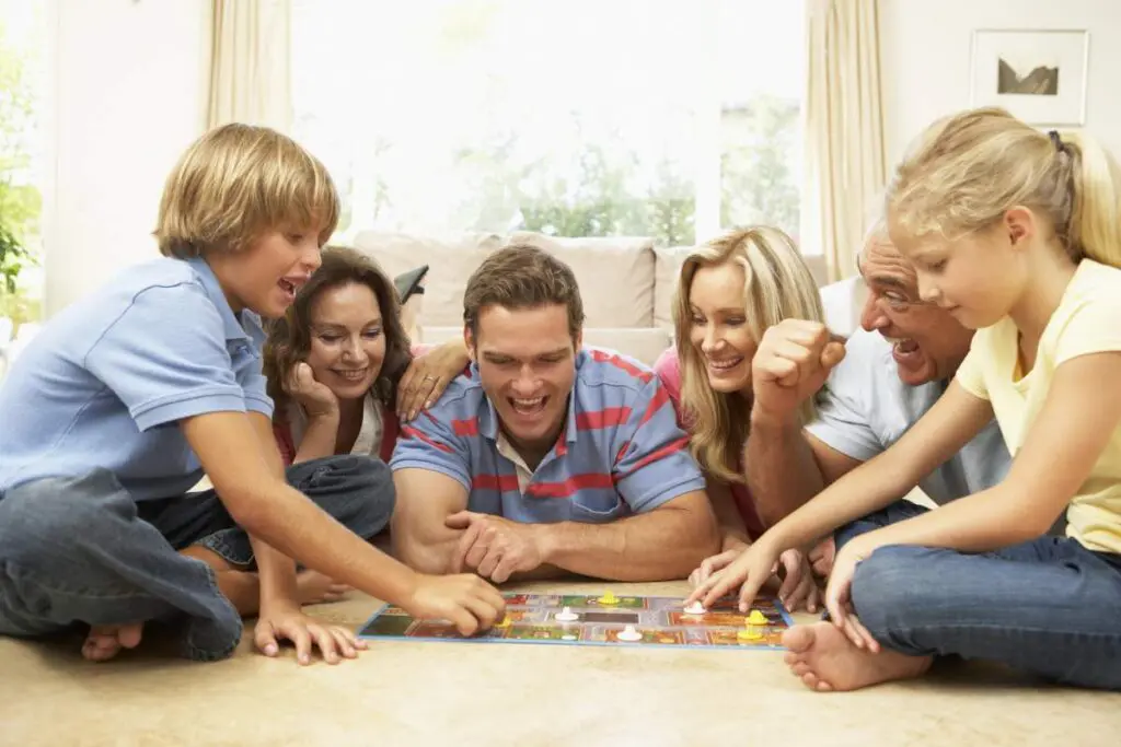 A group of four adults and two children are gathered around a board game, sitting on the floor of a living room, smiling and engaged in the game.