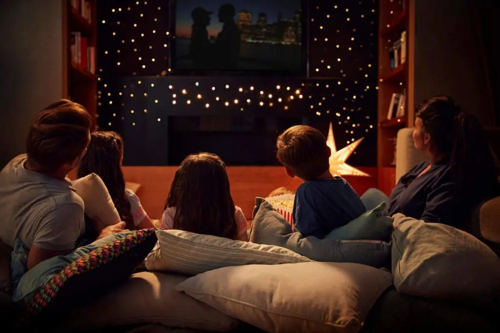A family sitting on a couch surrounded by cushions, watching a movie with fairy lights in the background.