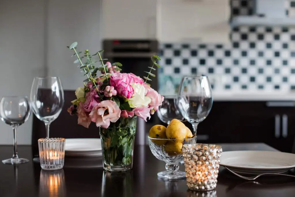 A dining table set with two empty wine glasses, a flower vase with pink flowers, a bowl of lemons.