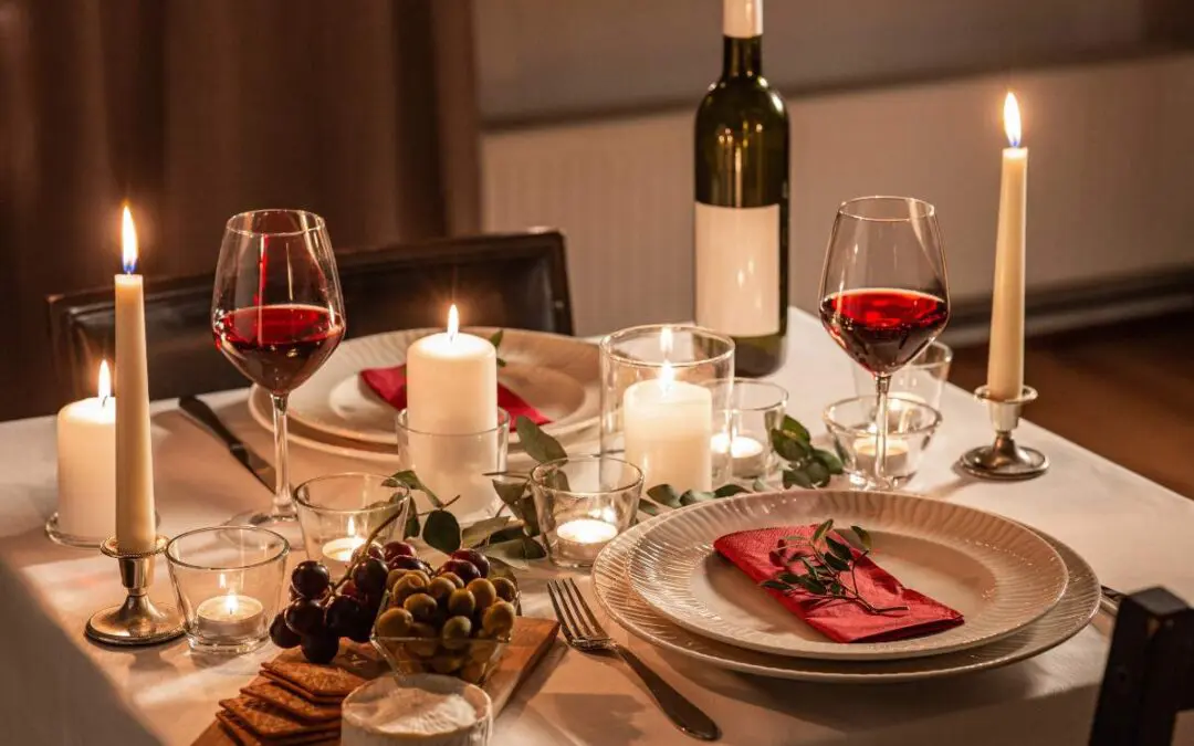 A beautifully set dinner table with red wine, candles, and elegant dishware, featuring grapes and cheese on a wooden board.