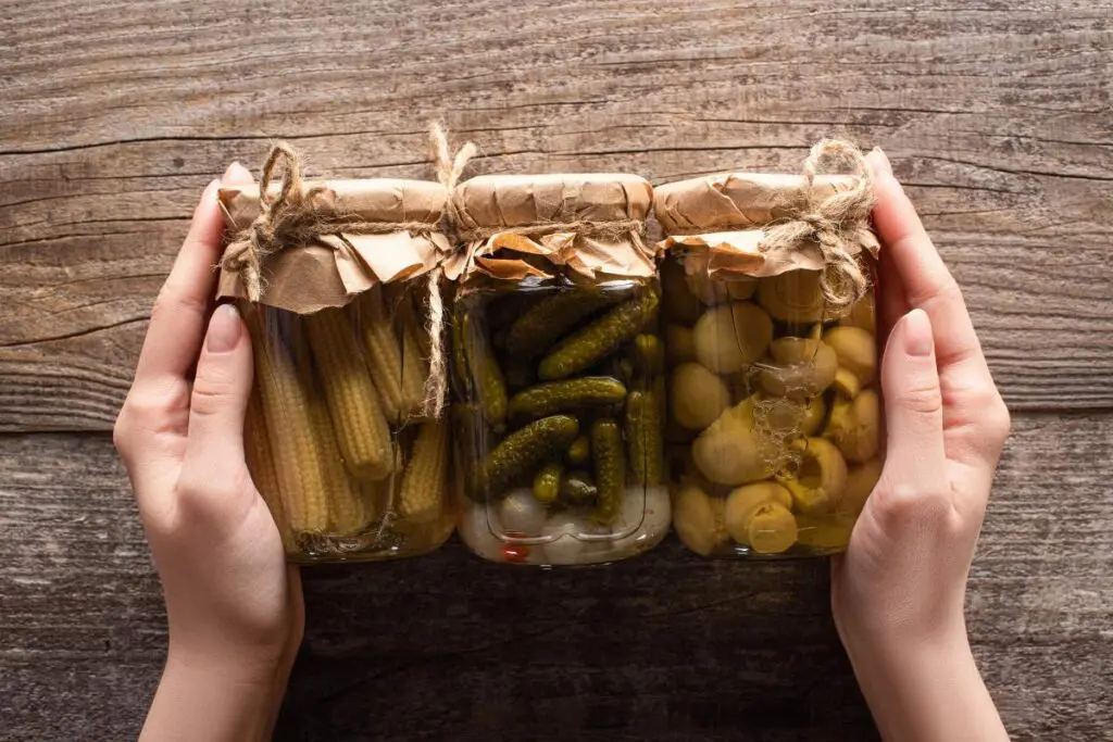 Three jars of preserved food held between two hands, each jar containing different types of pickled vegetables.