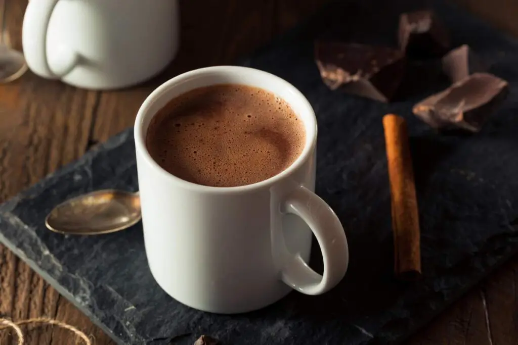 A white mug filled with hot chocolate, sitting on a slate surface, accompanied by pieces of dark chocolate and a cinnamon stick.