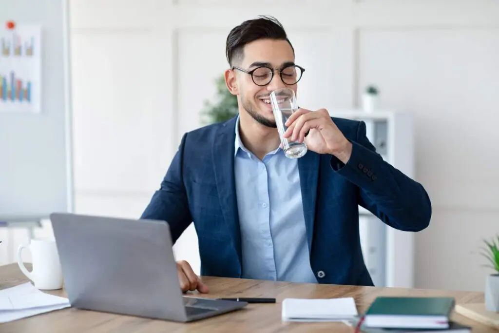 A man in a blue suit and glasses is smiling while drinking a glass of water at a desk with a laptop and notepad.