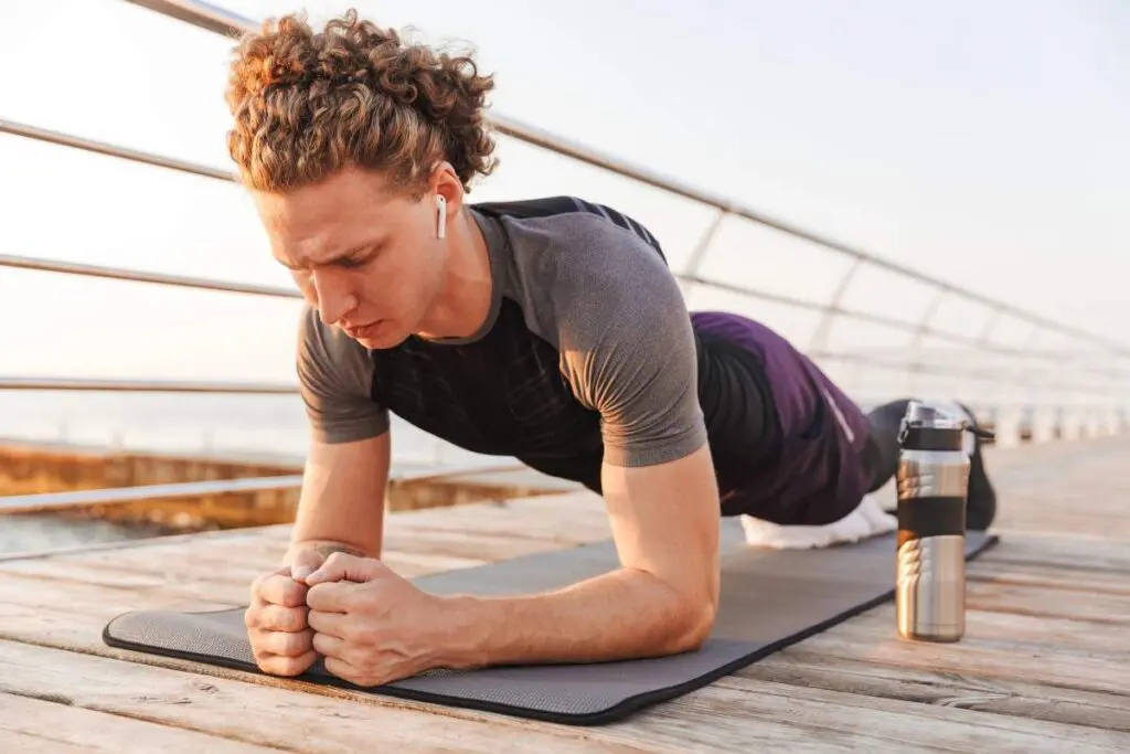 A man performs a plank exercise on a mat outdoors, wearing wireless earbuds and focused on his form. A water bottle is next to him.