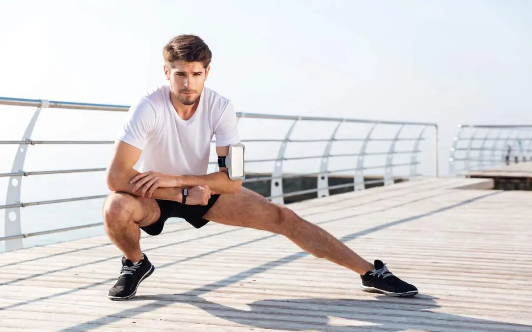 A young man in a white t-shirt and black shorts stretching on a wooden platform by the water.