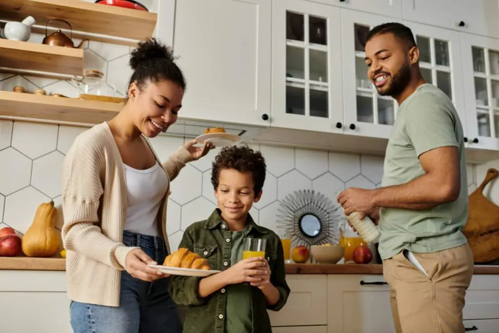 A woman and a man are smiling and interacting in a kitchen while a boy holds a glass of juice.