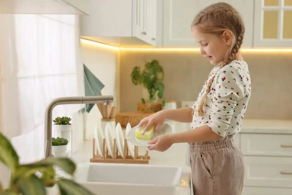 A young girl with braided hair is washing a plate at a kitchen sink, wearing a white sweater with floral patterns and beige pants.