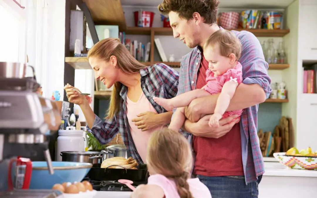 A family cooking together in a bright kitchen; a woman stirring a pot while holding a baby, and a man standing beside her. A young child is sitting on the floor facing away from them.