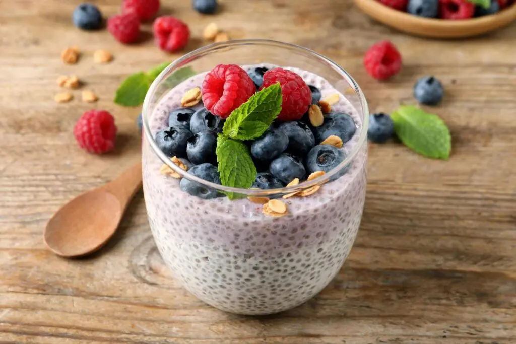 A glass of chia seed pudding topped with blueberries, raspberries, mint leaves, and granola on a wooden surface.