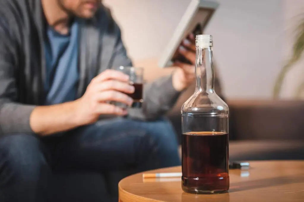 A person is holding a glass of dark liquid and a digital tablet, with a partially visible bottle of the same liquid on a coffee table.