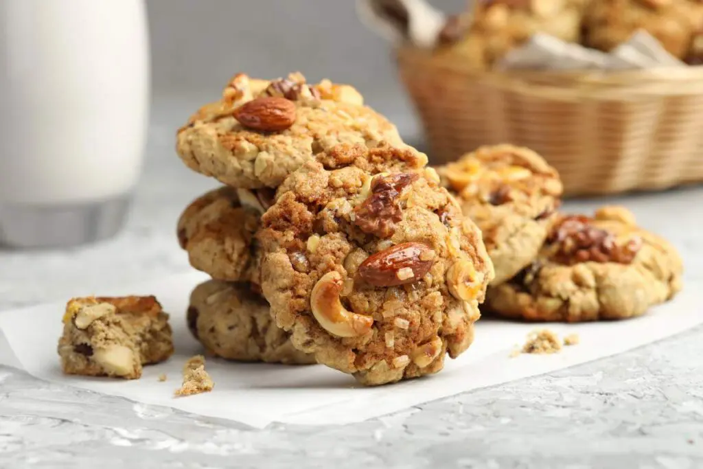 A stack of cookies with nuts on a white parchment paper, with more cookies in a wicker basket in the background and a glass of milk.